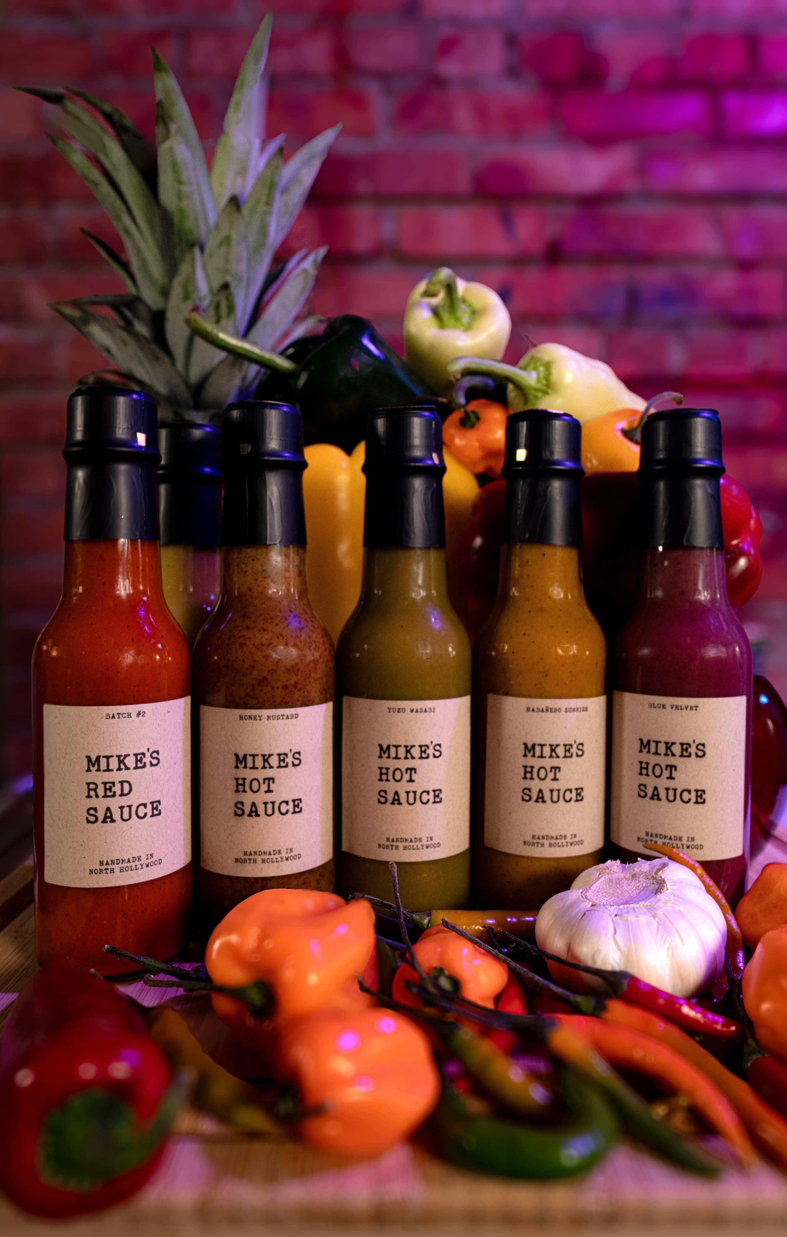 Six bottles from the Mike's Hot Sauce range arranged with peppers and a pineapple.